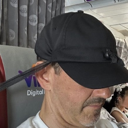 A man sleeping comfortably on an airplane, wearing a black Dreaming cap, which gently supports his head against the headrest.
